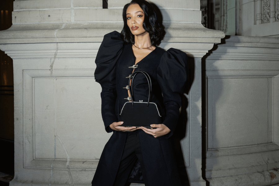 A woman in a black outfit with dramatic puffed sleeves holds a Giarité Cécile bag, a sculptural pearl kisslock bag, in front of a stone wall.