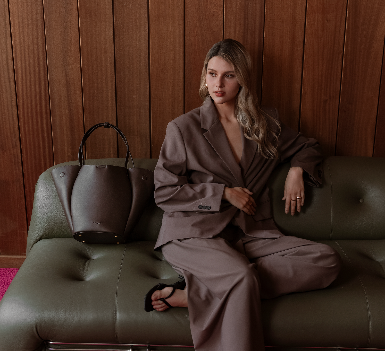 A woman with blonde hair wearing a brown pantsuit sits on a green couch next to a dark grey handbag, with a wooden wall behind her.
