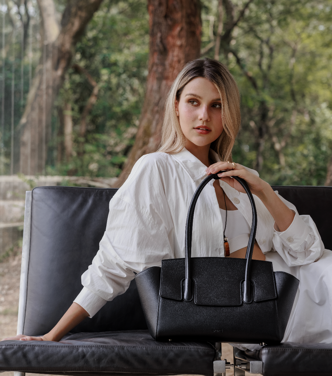 A woman in a white shirt sits on a dark couch, holding a black Giarité Valyn Tote bag. Trees and foliage are visible in the blurred background.