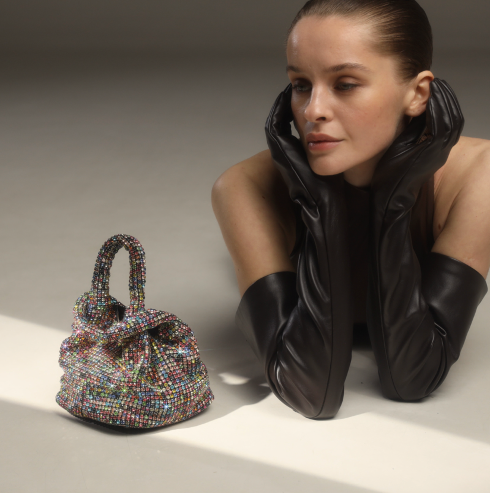 A woman with pulled-back hair and long black gloves rests her chin on her hands, looking towards a Giarité Brilly Hue multicolour rhinestone bucket bag positioned in the foreground.