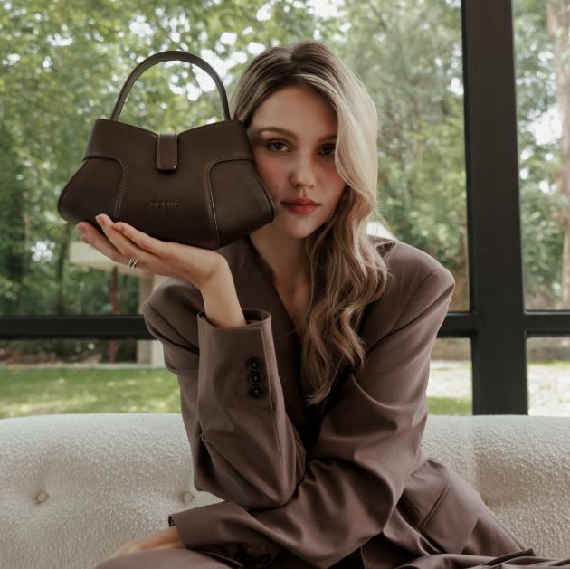 A woman with long, light brown hair sits indoors, holding up a dark brown Giarité Paphia handbag with her left hand, showcasing it prominently.