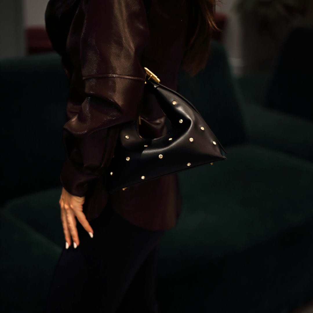 A black, structured vegan leather shoulder bag with gold-tone stud embellishments and a gold strap detail, held by a person wearing a dark brown jacket and black pants, against a blurred dark background.