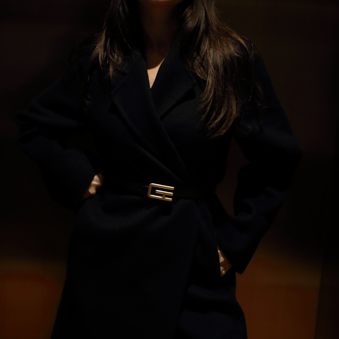The image displays a woman wearing a black coat cinched at the waist with a belt featuring a prominent gold-toned buckle, which is identified as the ""GTE Belt"" from Giarité