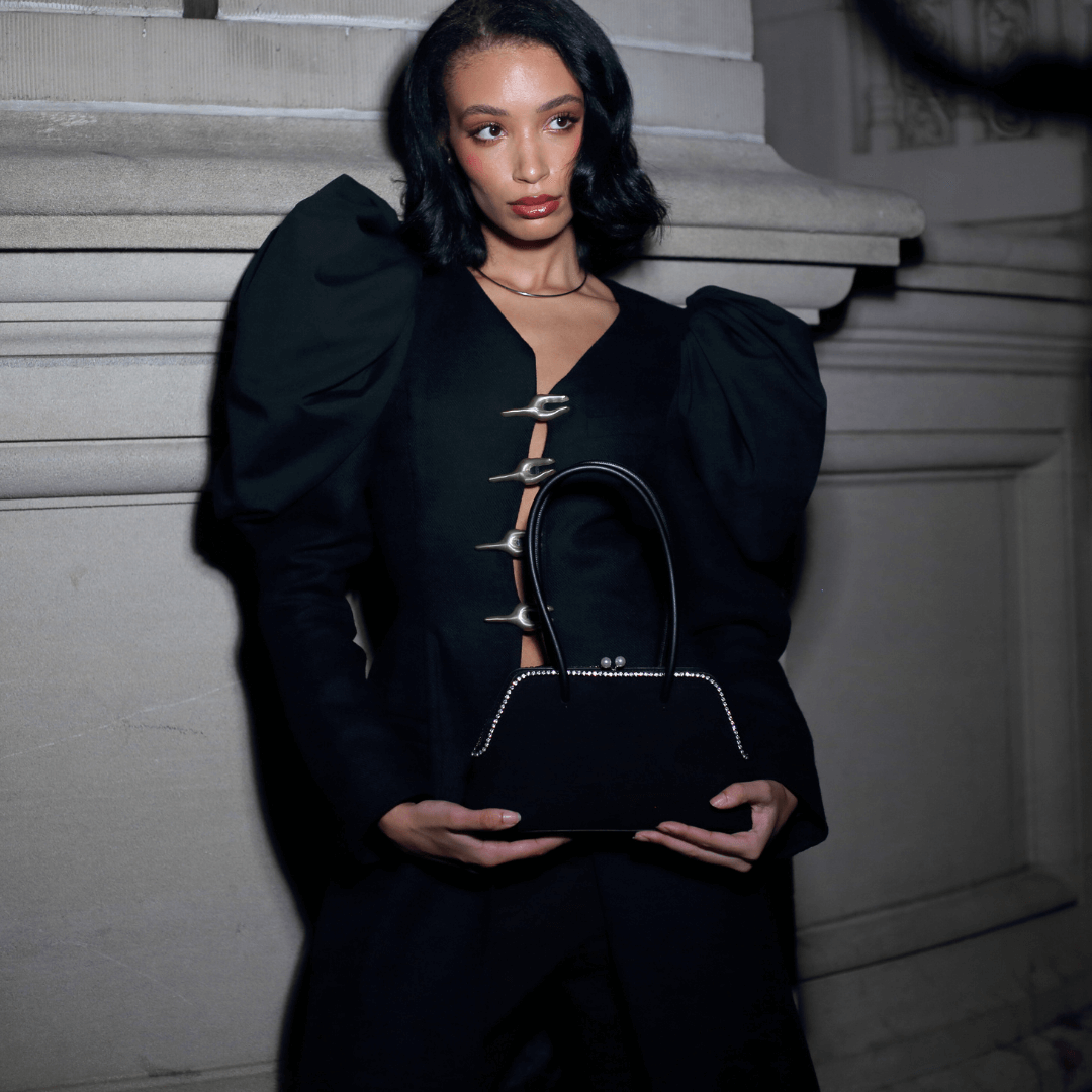 A black structured handbag, handcrafted with hand-set luminous pearls across a structured, sculptural base, held by a model wearing a black top with puffed sleeves and silver embellishments
