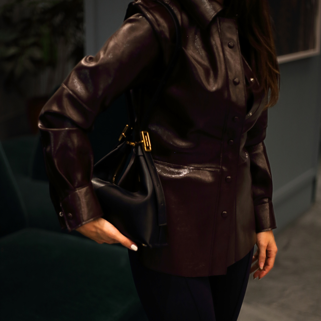 A person wearing a dark, possibly leather, shirt holds a black Jules Grande handbag, which appears to be a structured vegan hobo shoulder bag.