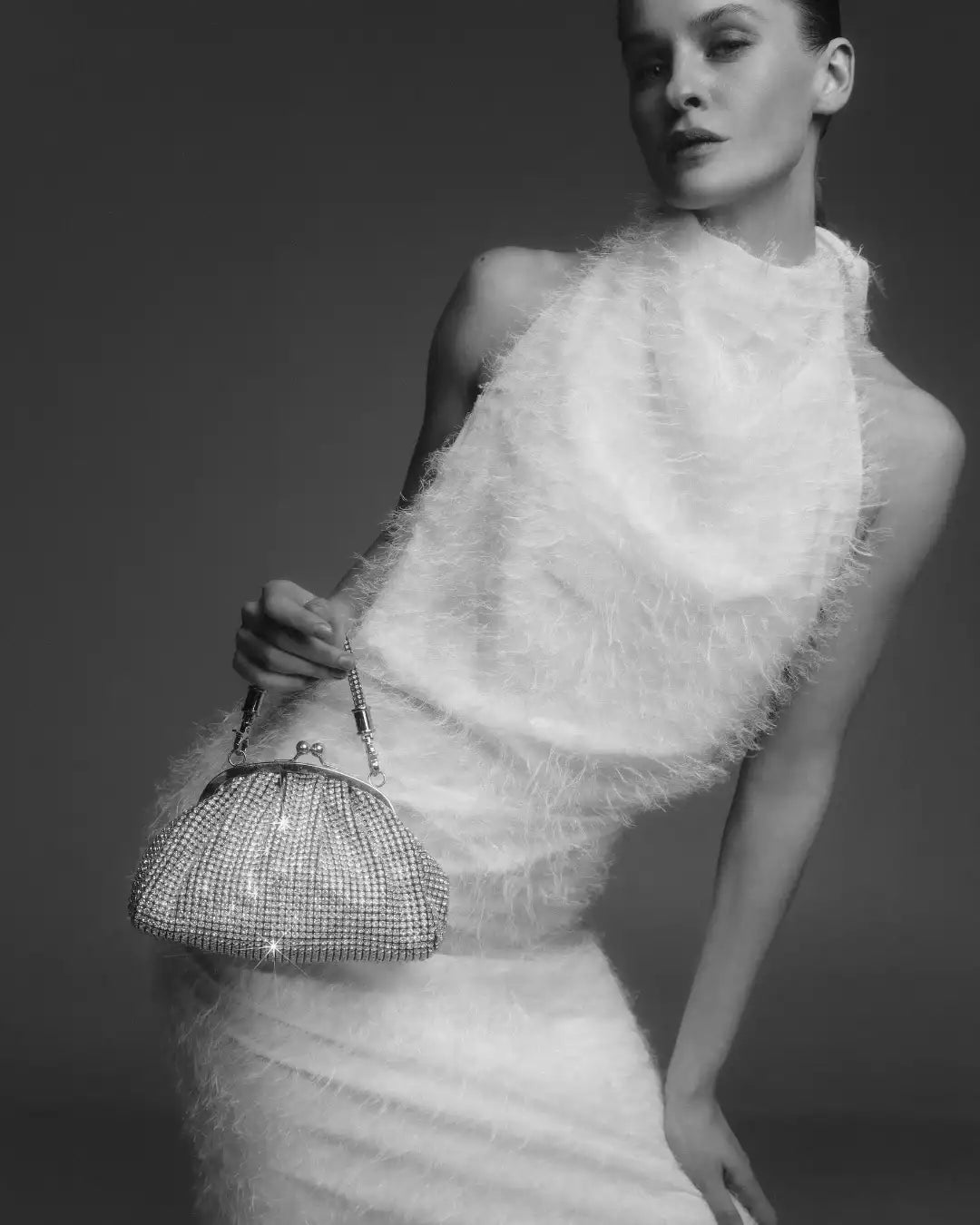 A black and white image of a model in a white, textured dress, holding Giarite's silver Lima handbag.