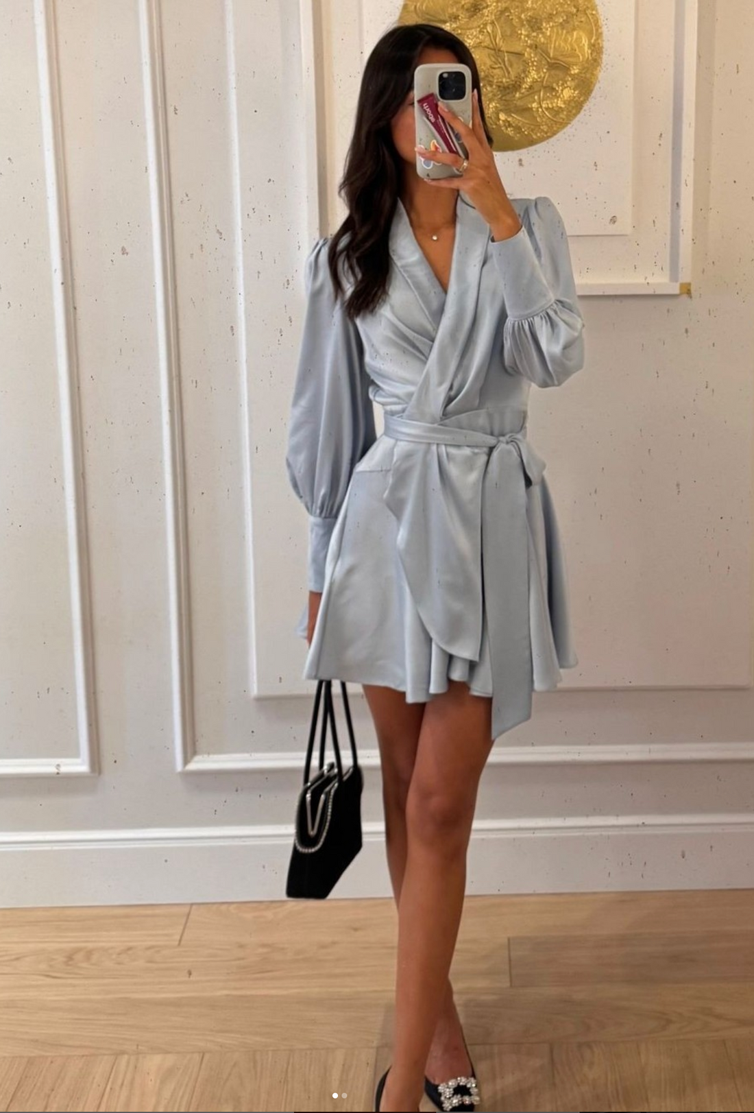 A woman with long dark hair, wearing a light blue, long-sleeved, asymmetrical wrap mini-dress and dark embellished flat shoes, stands indoors holding a black handbag. She is taking a mirror selfie, and part of a decorative gold object is visible in the reflection behind her.