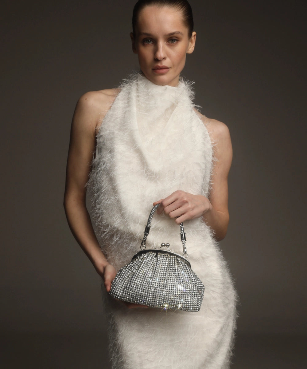 A woman in a white, textured, high-neck dress holds a silver, rhinestone-embellished clutch bag with a chain strap in front of a neutral background.