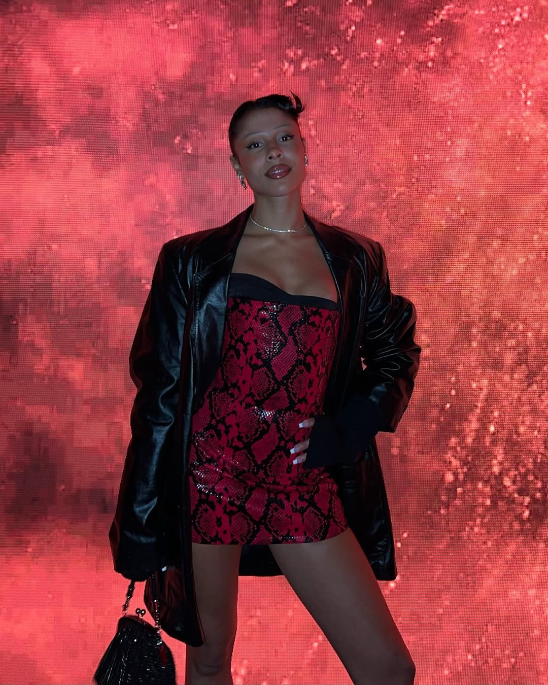 A woman with dark hair pulled back, wearing a black leather jacket over a red and black snakeskin-patterned strapless dress, stands in front of a vibrant red, textured background. She is holding a black purse and looking directly at the camera.