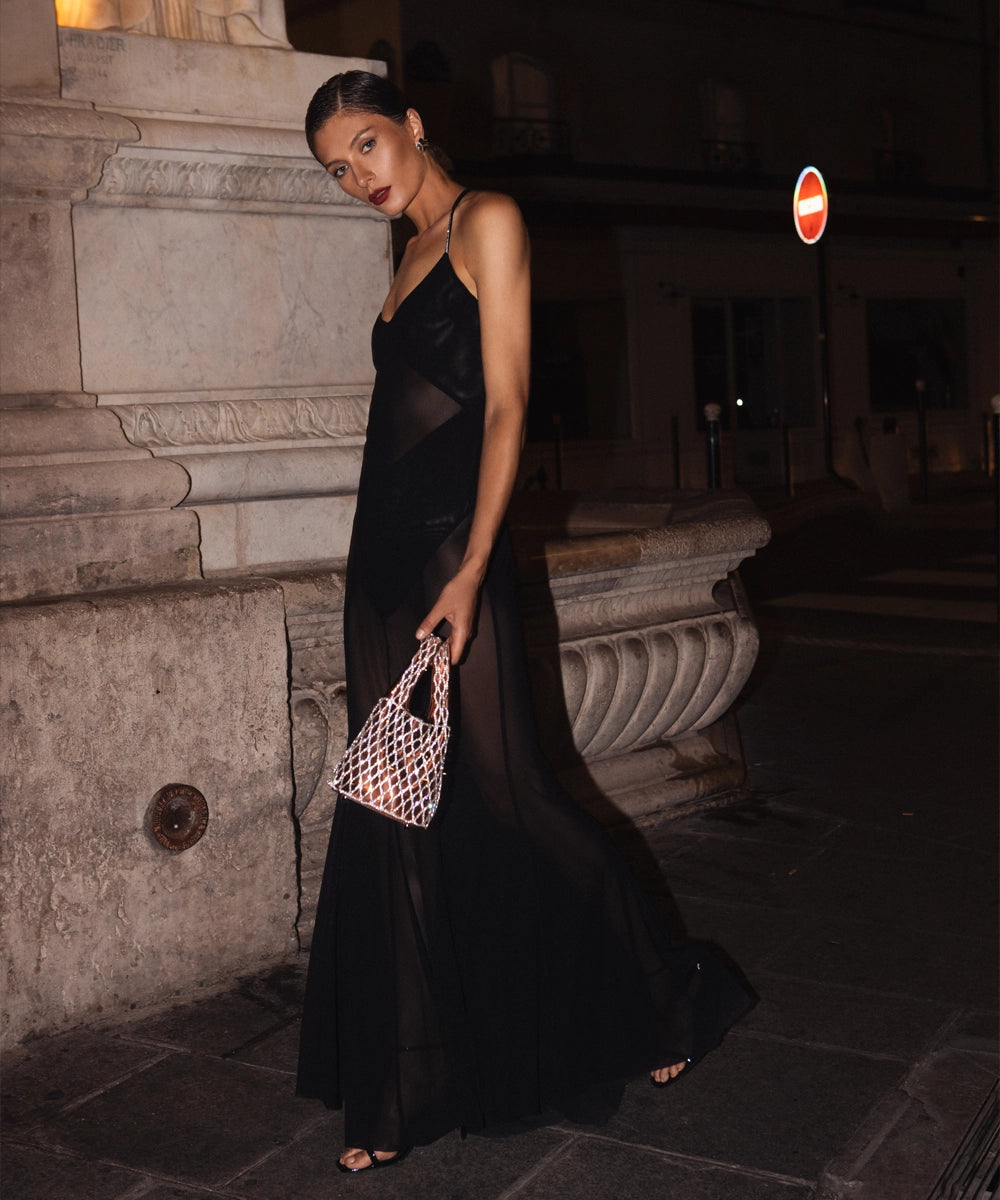 A woman in a sheer black maxi dress with a plunging neckline and thigh-high slit holds a sparkling, iridescent handbag while standing on a stone walkway at night.