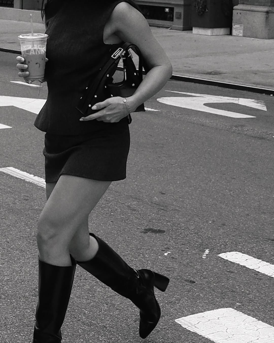 A black and white photo shows a person from the waist down, walking across a street. They are wearing a dark dress or top and skirt, knee-high boots, and holding a studded shoulder bag in one hand and a clear plastic cup with a drink in the other. White road markings are visible on the asphalt.