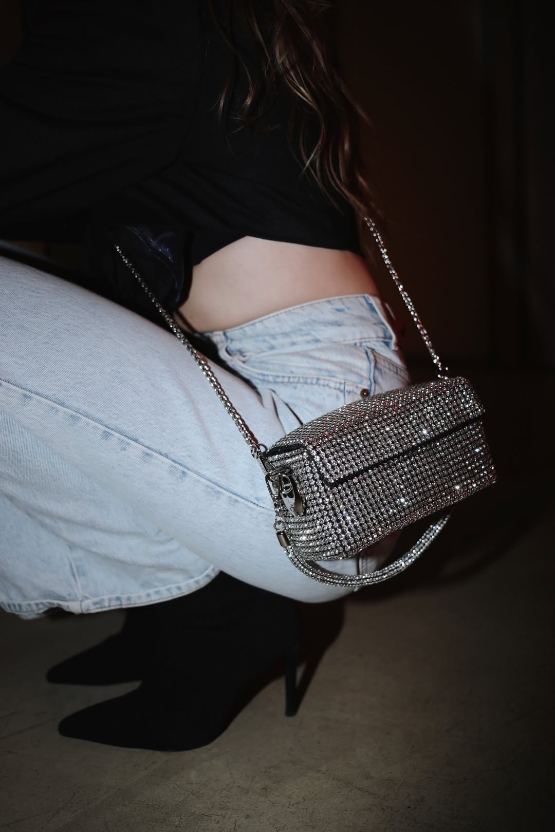 A person in light-wash jeans and black heels holds a shimmering silver rhinestone box clutch bag with a chain strap.