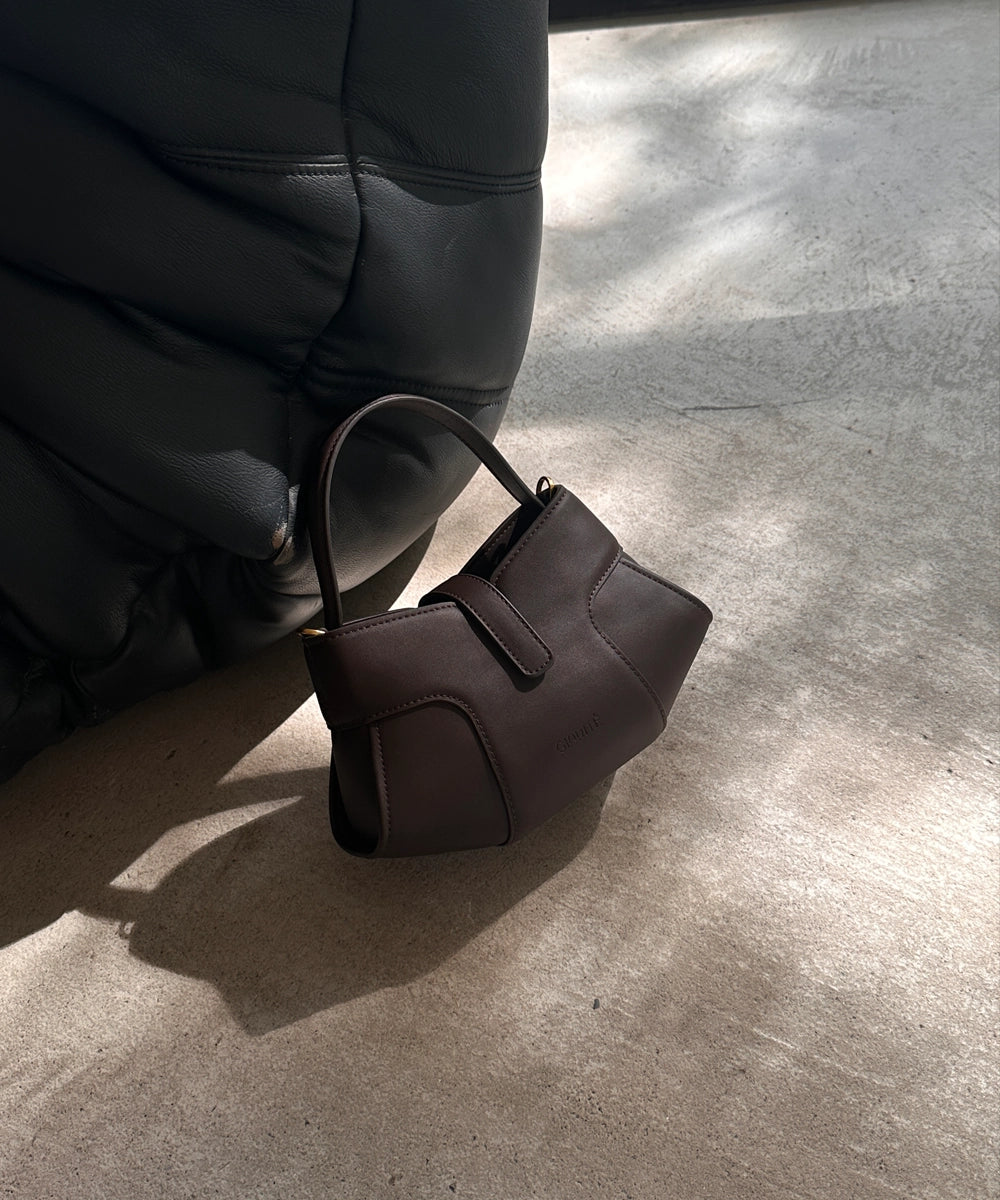 The image depicts a dark brown, structured handbag with a top handle, resting on a light-colored, possibly concrete or stone, floor. A dark, possibly leather or upholstered, object, which appears to be part of a chair or sofa, is partially visible in the background, creating a shadow on the floor. The lighting suggests an indoor setting with natural light coming from a window or opening, casting distinct shadows.