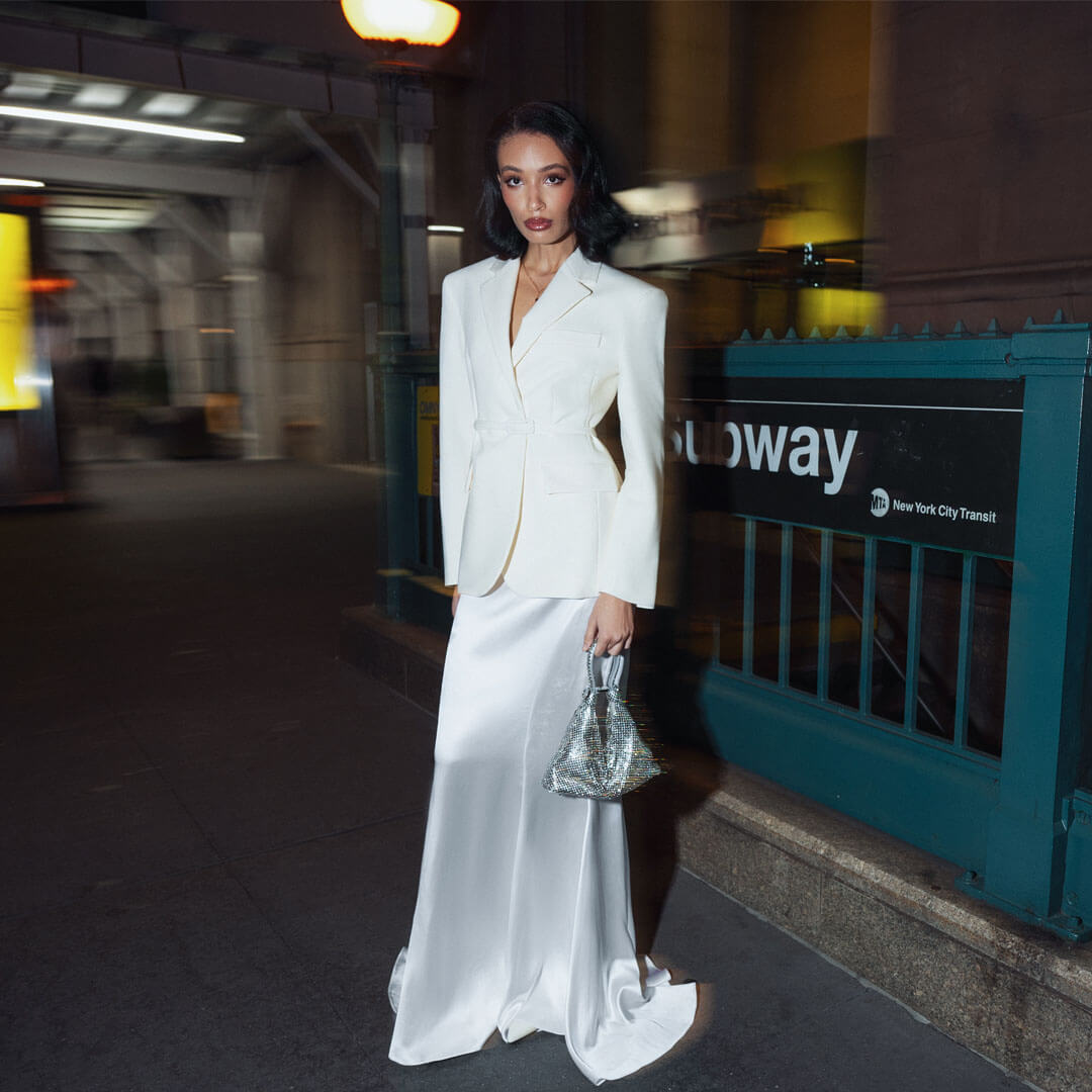 A woman in a white blazer and a long white satin skirt stands in a subway station, holding a Giarité's Jules Mirage Silver handbag.
