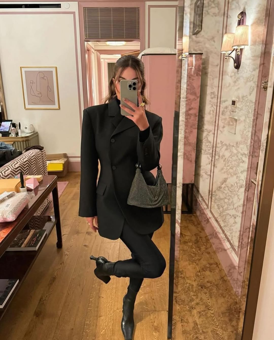 A woman wearing a black blazer, black pants, and black boots stands in a room with a mirror, taking a selfie while holding a small gray textured handbag. The room features a patterned chair, a wooden table with various items, and a wall with a framed abstract artwork and a wall sconce.
