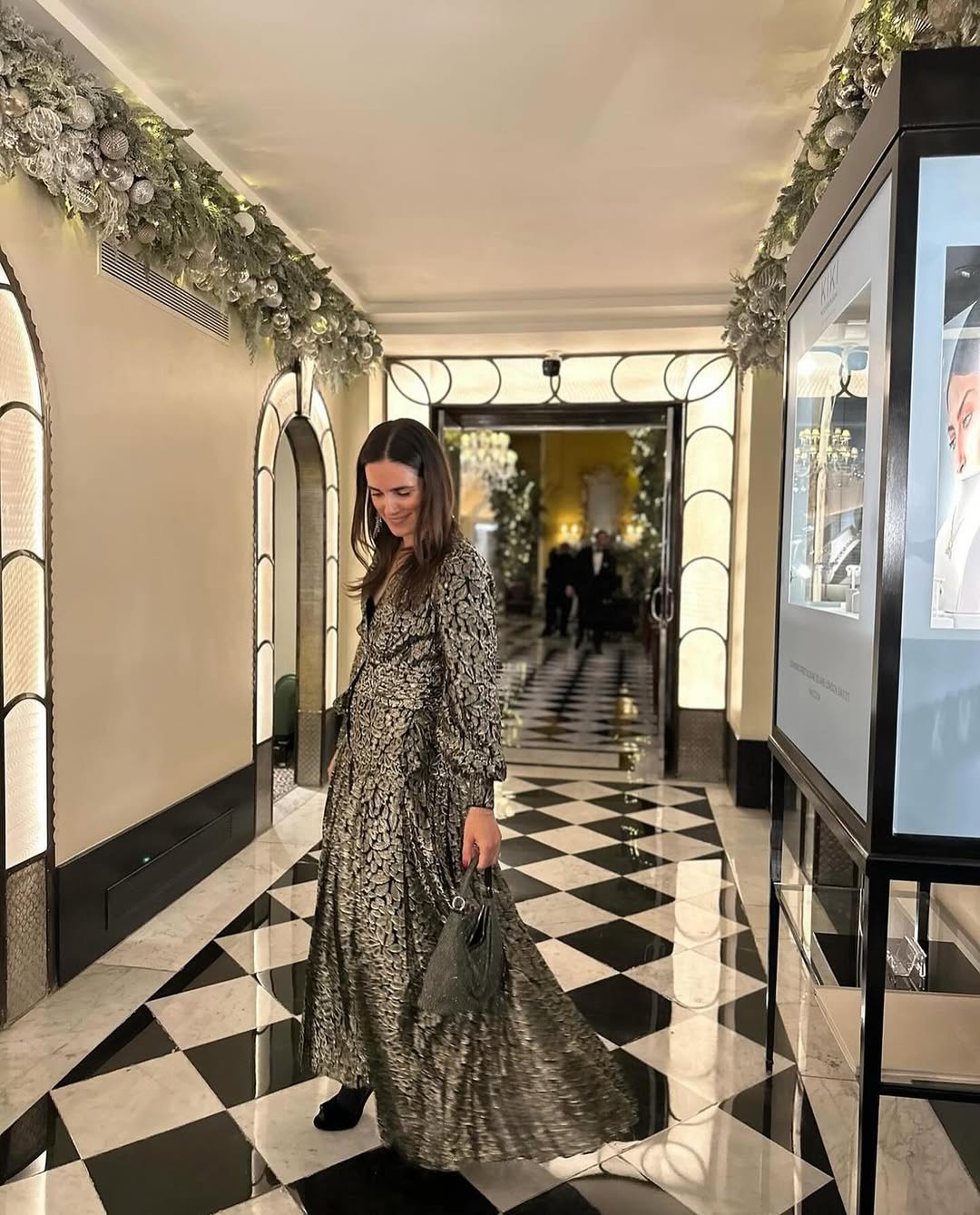 A woman in a long, patterned dress and dark shoes walks down a hallway with a black and white checkered floor, carrying a dark handbag. The hallway is decorated with festive garland along the ceiling and features illuminated display cases on the right and archways on the left. In the background, a brightly lit room with more people is visible through an open doorway.