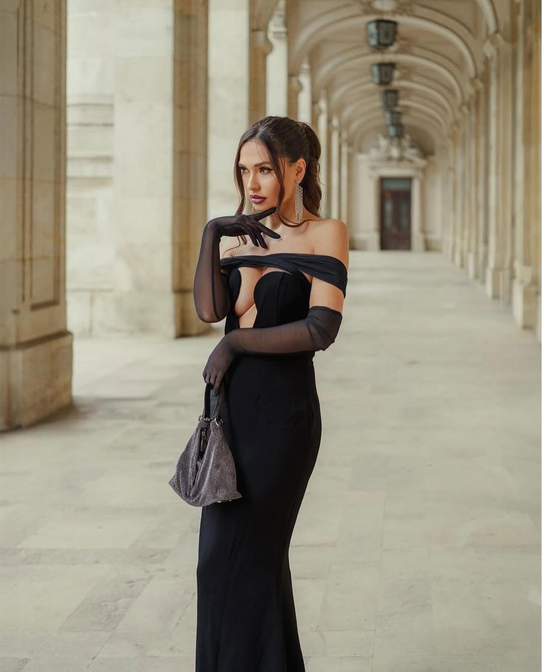 A woman in a long, off-the-shoulder black dress with a deep V-neckline and sheer black gloves stands in a grand hallway with arched ceilings and columns. She holds a grey, textured handbag and looks to her right, with her left hand near her chin.