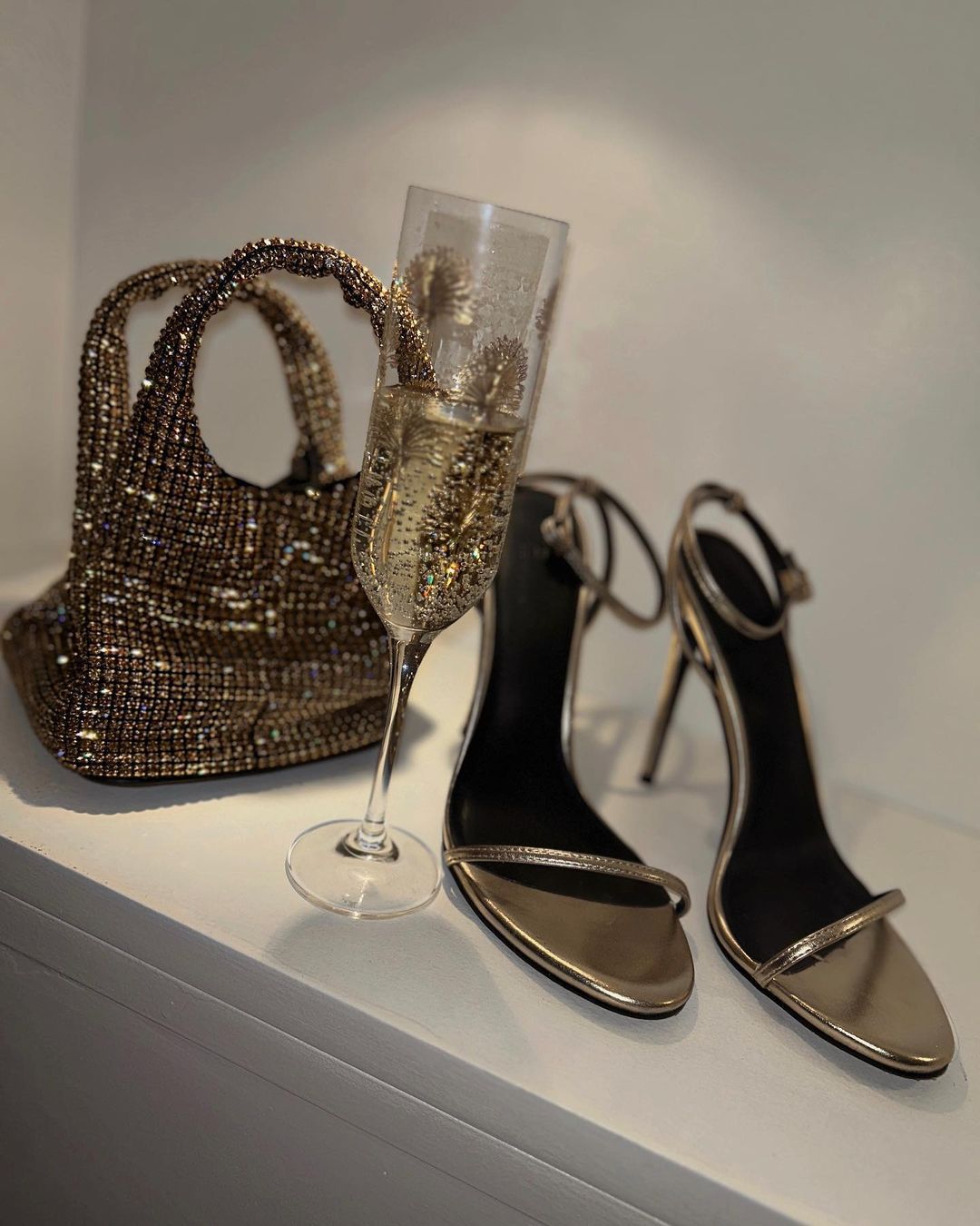 A sparkling gold handbag, a champagne flute filled with bubbly, and a pair of gold high-heeled sandals with delicate straps, all arranged on a white surface against a light background.