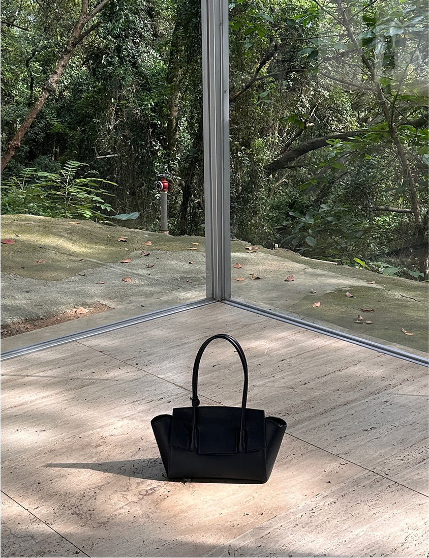 A black handbag sits on a light-colored tiled floor in front of a large window looking out onto a lush green forest or garden. The window frame is visible, and the natural light casts a shadow from the handbag on the floor.