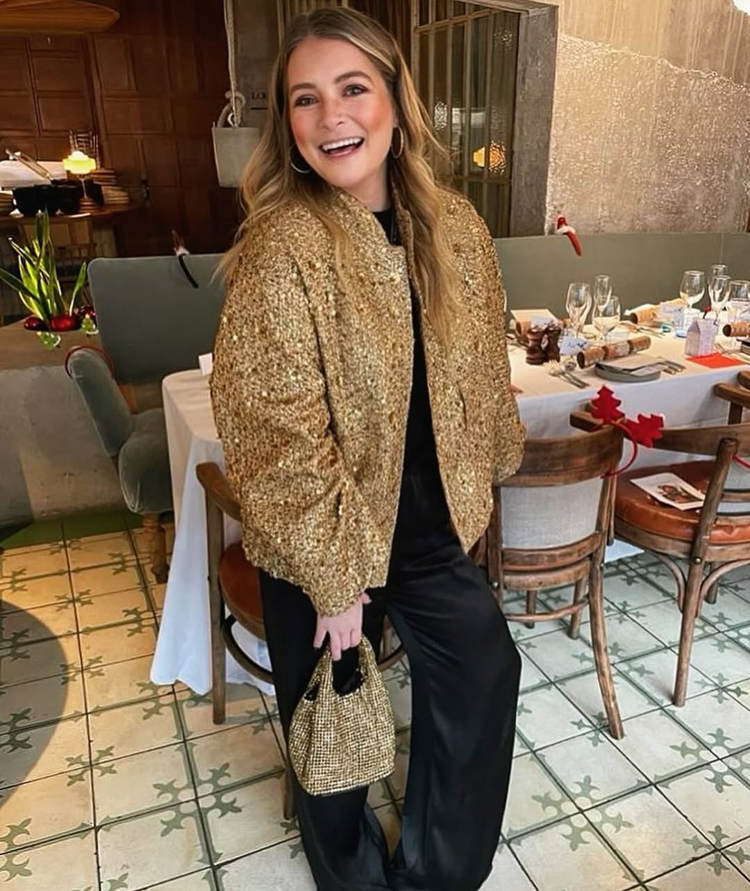A smiling woman with long blonde hair and hoop earrings stands in a restaurant, wearing a sparkly gold bomber jacket, black pants, and holding a matching small gold sequined handbag. In the background, there are dining tables set with white tablecloths and chairs, with a plant visible on a nearby table.