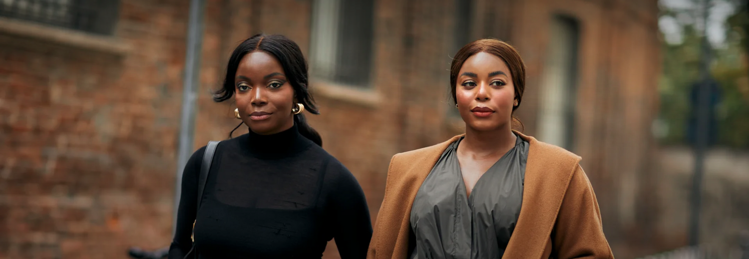 Two Black women, one in a black top and the other in a brown coat over a grey top, stand side-by-side outdoors with a brick building in the background.