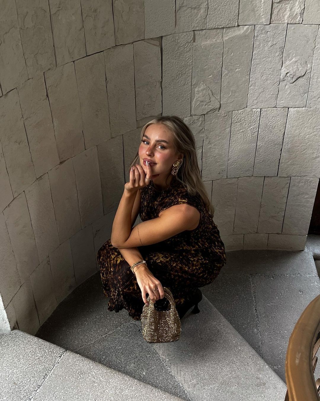 A young woman with long blonde hair, wearing a dark leopard print dress and earrings, crouches on a grey stone staircase, holding a small, sparkly gold handbag. She is looking at the camera with a slight smile, resting her chin on her hand. The background is a textured grey stone wall.
