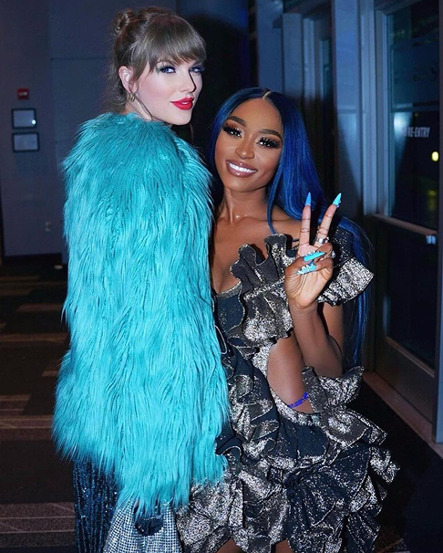 Taylor Swift in a teal fuzzy coat stands next to Sonyae Elise, who has blue hair and is wearing a ruffled black and gold dress, flashing a peace sign.