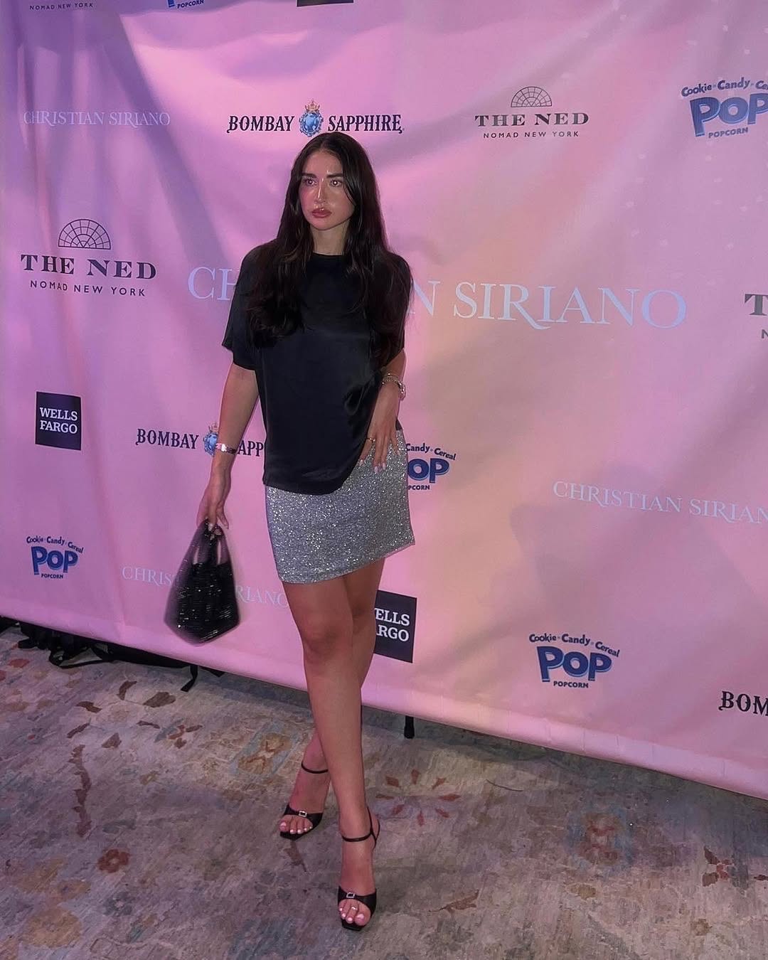 A woman with long dark hair, wearing a black top, a sparkly silver mini skirt, and black high-heeled sandals, poses on a red carpet in front of a pink backdrop with various brand logos like Christian Siriano, Bombay Sapphire, The Ned, Wells Fargo, and Pop Popcorn. She is holding a black handbag in her left hand and looking directly at the camera.