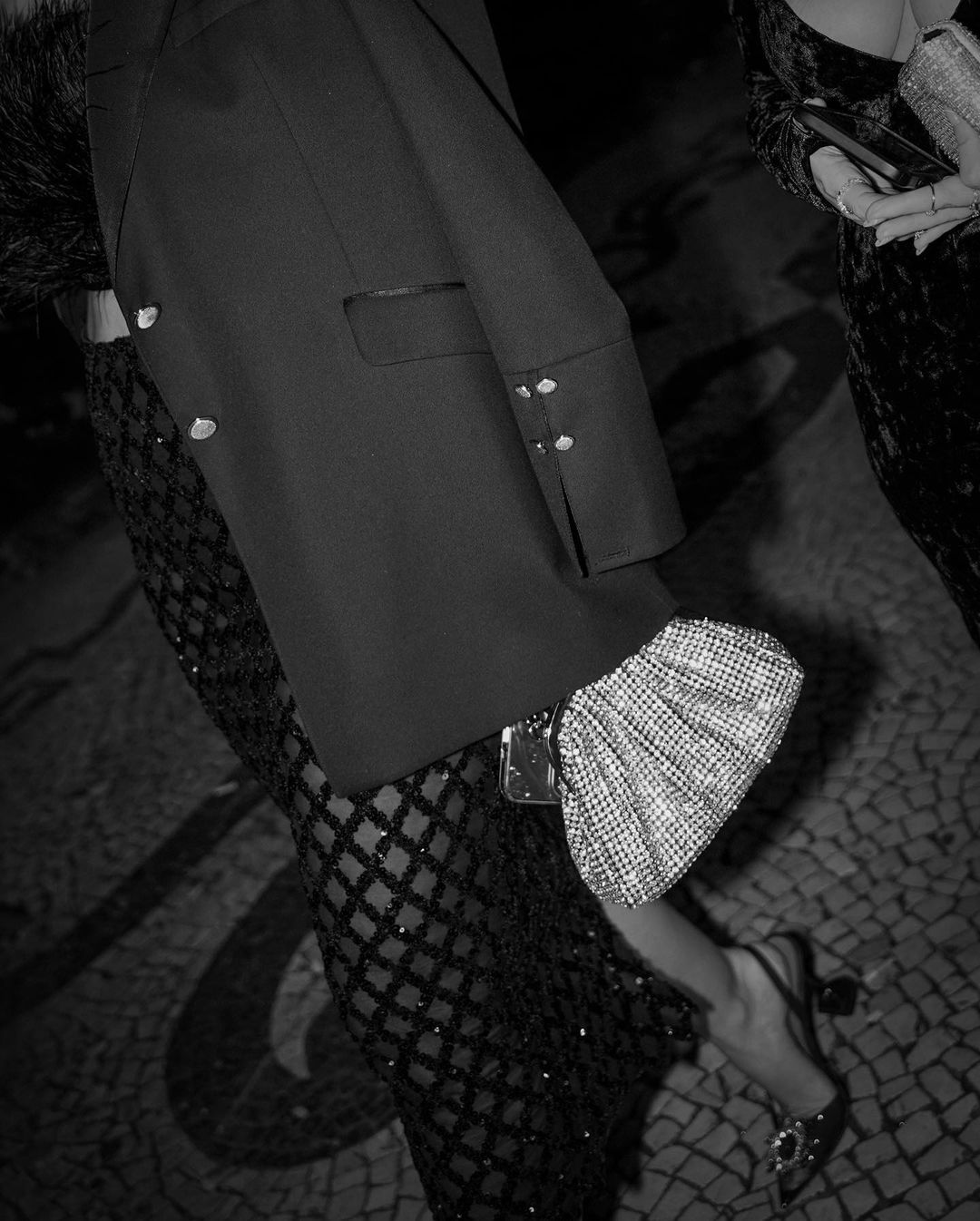 A black and white, low-angle shot captures a person from the waist down, walking on a patterned surface. The person wears a dark, long-sleeved top or jacket with button details, a shimmering, dark, patterned skirt or dress, and high-heeled shoes with a buckle detail. They are holding a sparkling, textured clutch bag. In the upper right corner, another person's arm and a textured clutch are partially visible.