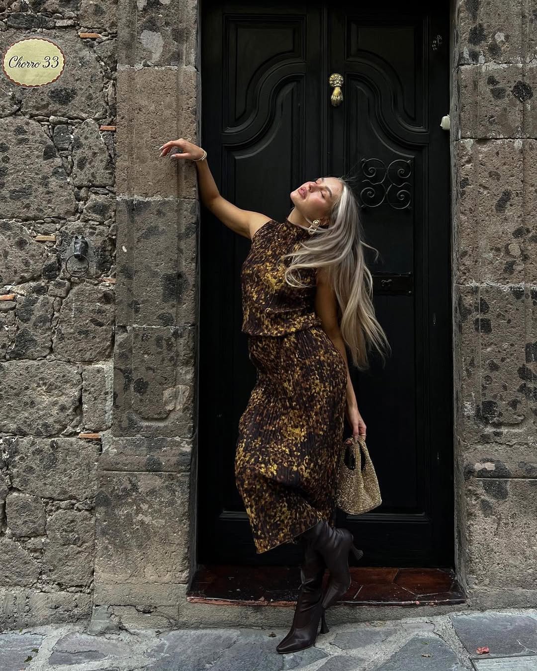 A woman with long blonde hair, wearing a leopard print top and matching midi skirt, dark boots, and carrying a small woven bag, poses with her head tilted back and arm raised against a textured stone wall next to a dark wooden door with a brass knocker. A small plaque on the wall reads Chorro 33.