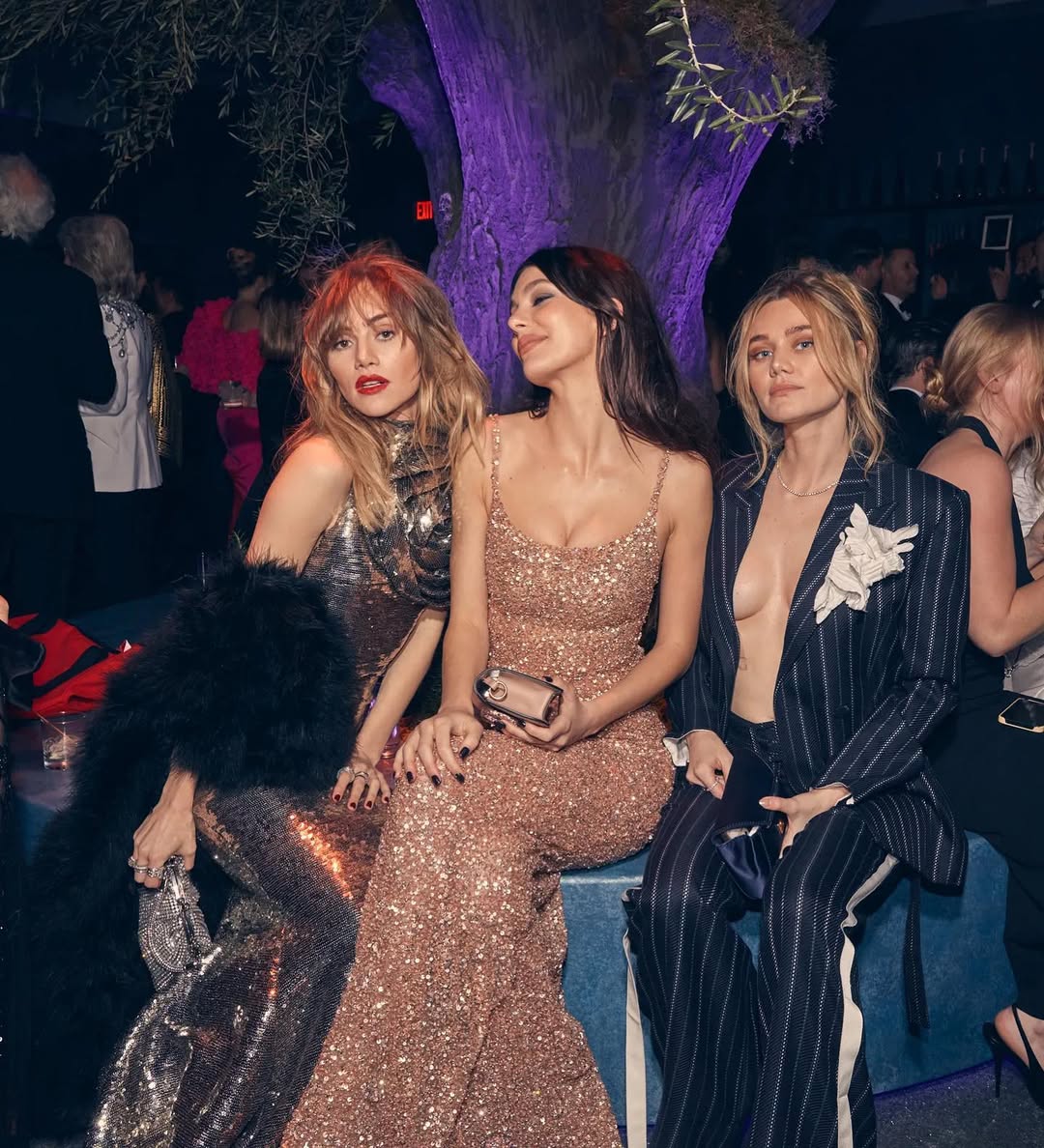 Three women, Suki Waterhouse, Camila Morrone, and Immy Waterhouse, posing at the Vanity Fair Oscars after-party. Suki Waterhouse is on the left in a shimmering gold gown with a black fur stole, Camila Morrone is in the center in a rose gold sequined dress, and Immy Waterhouse is on the right in a pinstriped suit jacket with a floral embellishment.