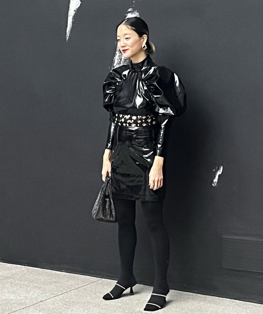 A woman with her hair pulled back, wearing a shiny black vinyl mini-dress with dramatic puffed sleeves, a high neck, and a crystal-embellished cutout waist, paired with black tights and two-tone black and white heels, stands against a dark gray wall with white markings.