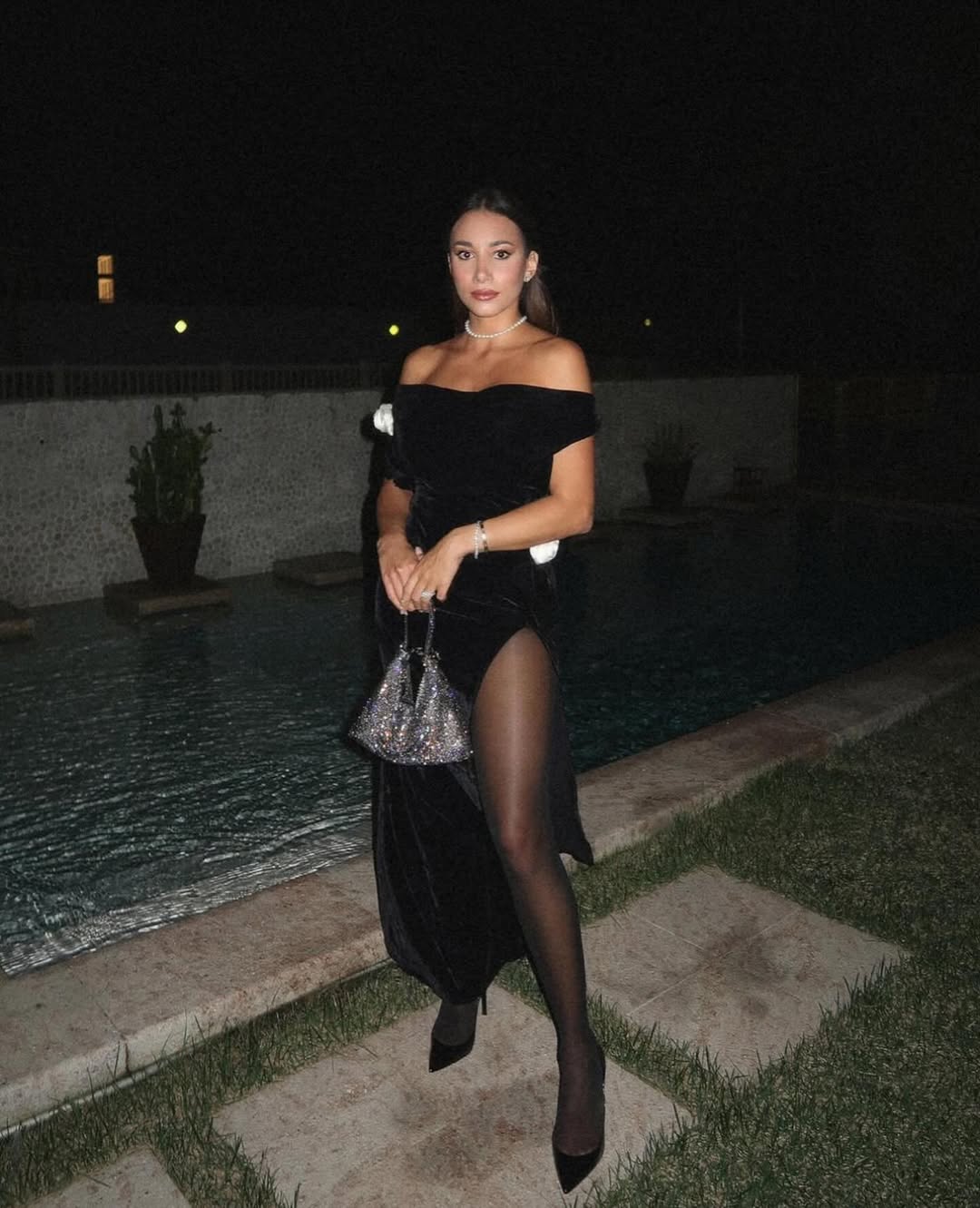 A woman wearing a black off-the-shoulder gown with a high slit, black sheer stockings, and black heels stands beside a swimming pool at night. She is holding a small, sparkling handbag and wearing a choker necklace and a bracelet. The background shows a dimly lit outdoor area with the pool and some potted plants.