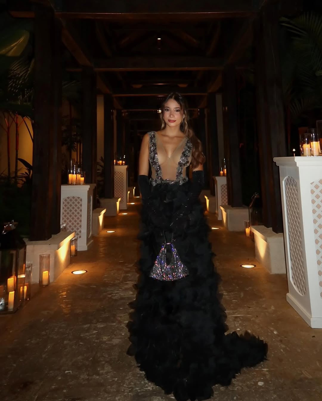 A woman in a black, ruffled gown with a sheer, embellished bodice and long black gloves stands in a dimly lit, candle-adorned hallway or pathway at night, holding a sparkling clutch bag.