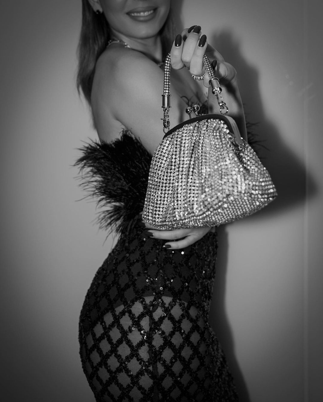 A black and white, close-up shot of a woman in a glamorous, sequined dress with feather details, holding a sparkling silver diamante handbag with a chain strap. She is smiling, and the focus is on her torso and the handbag, showcasing a sophisticated and elegant style.