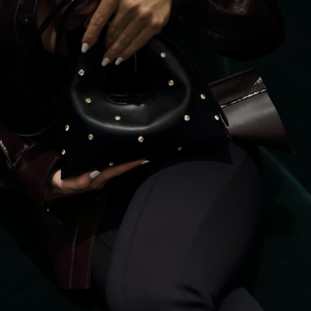 A black handbag with a distinctive curved shape, adorned with scattered clear rhinestones or studs across its surface, held by hands with white manicured nails, displayed on what appears to be a monitor screen.