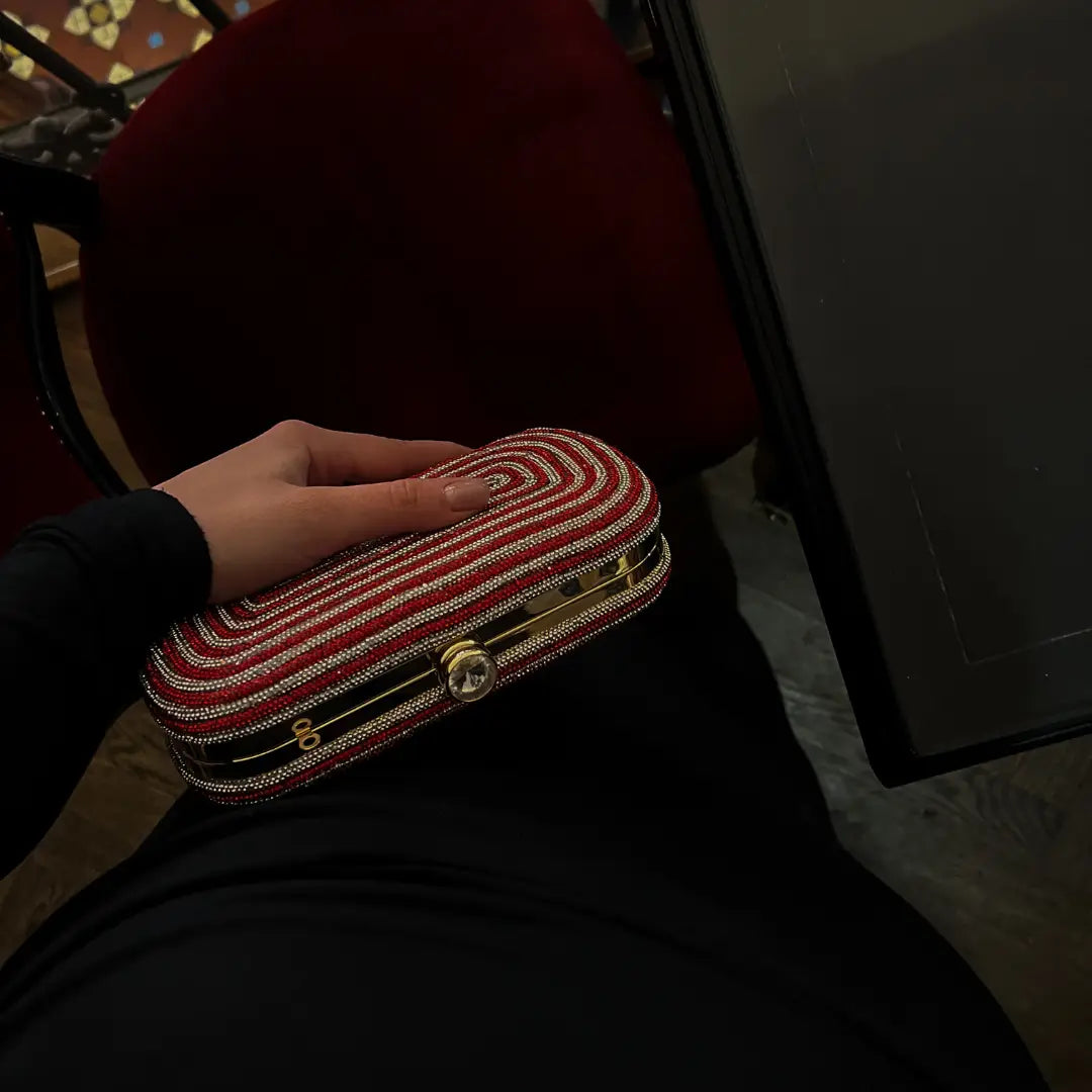 A person in a black long-sleeved top holds a Giarité Crystal Minaudière clutch bag, which is red and white with a gold-tone frame and push-lock closure, against a dark background with a red chair visible.