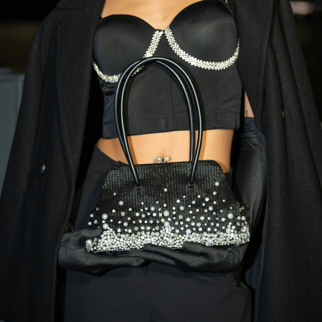 A person in a black outfit, including a black satin bralette with crystal embellishments and long black gloves, holds Giarité's Cécile Pearlfall handbag, a black beaded evening purse adorned with cascading pearl strands and crystal beads. 