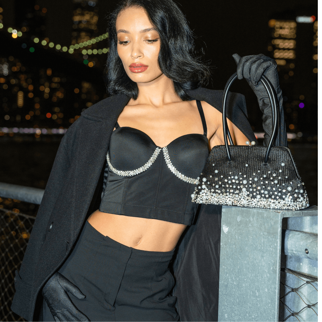 A woman in a black outfit, including a rhinestone-embellished bustier top and gloves, holds a Giarité's Cécile Pearlfall handbag, which is a black beaded evening purse with cascading pearl strands and crystal beads, against a blurry nighttime city backdrop. 