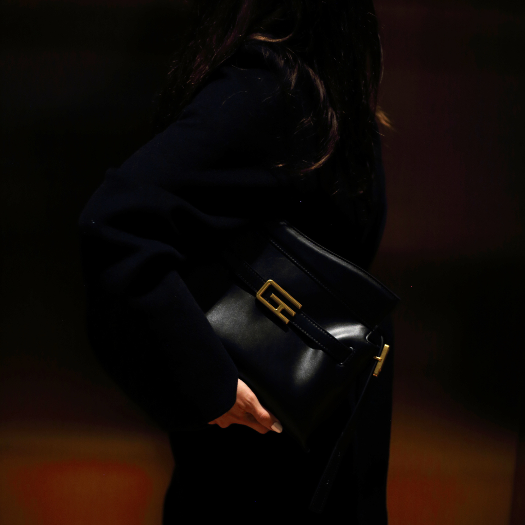  A person in dark clothing carries a black Giarité Trapezio handbag with a prominent gold-toned buckle on its flap, held under their arm against a dark, subtly lit background, highlighting the bag's structure. 