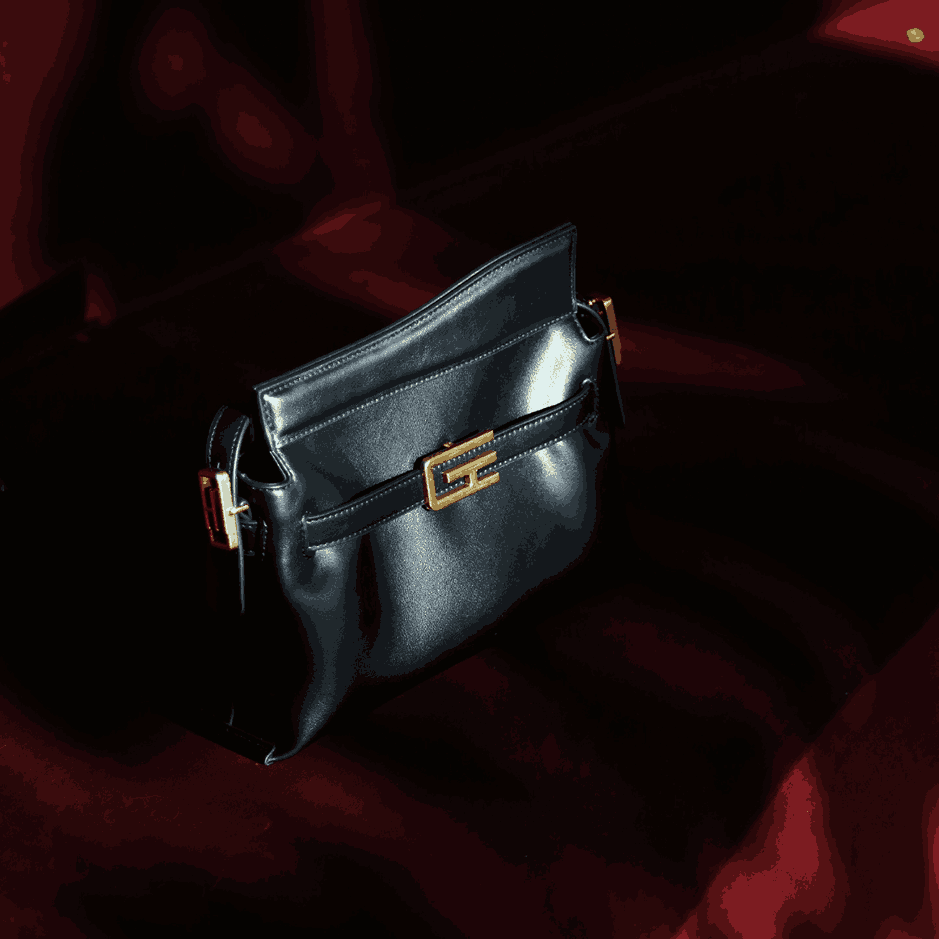 A black Giarité G-TRAPEZIO vegan leather shoulder bag with gold-toned hardware, featuring a distinctive trapezoid shape and a flap closure with a G-shaped buckle, resting on a dark red velvet surface.
