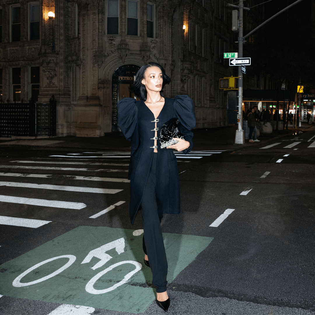 A woman in a dark, structured coat with prominent shoulders and dark pants walks across a city street at night, holding Giarite's Pearlfall Limited Edition handbag.