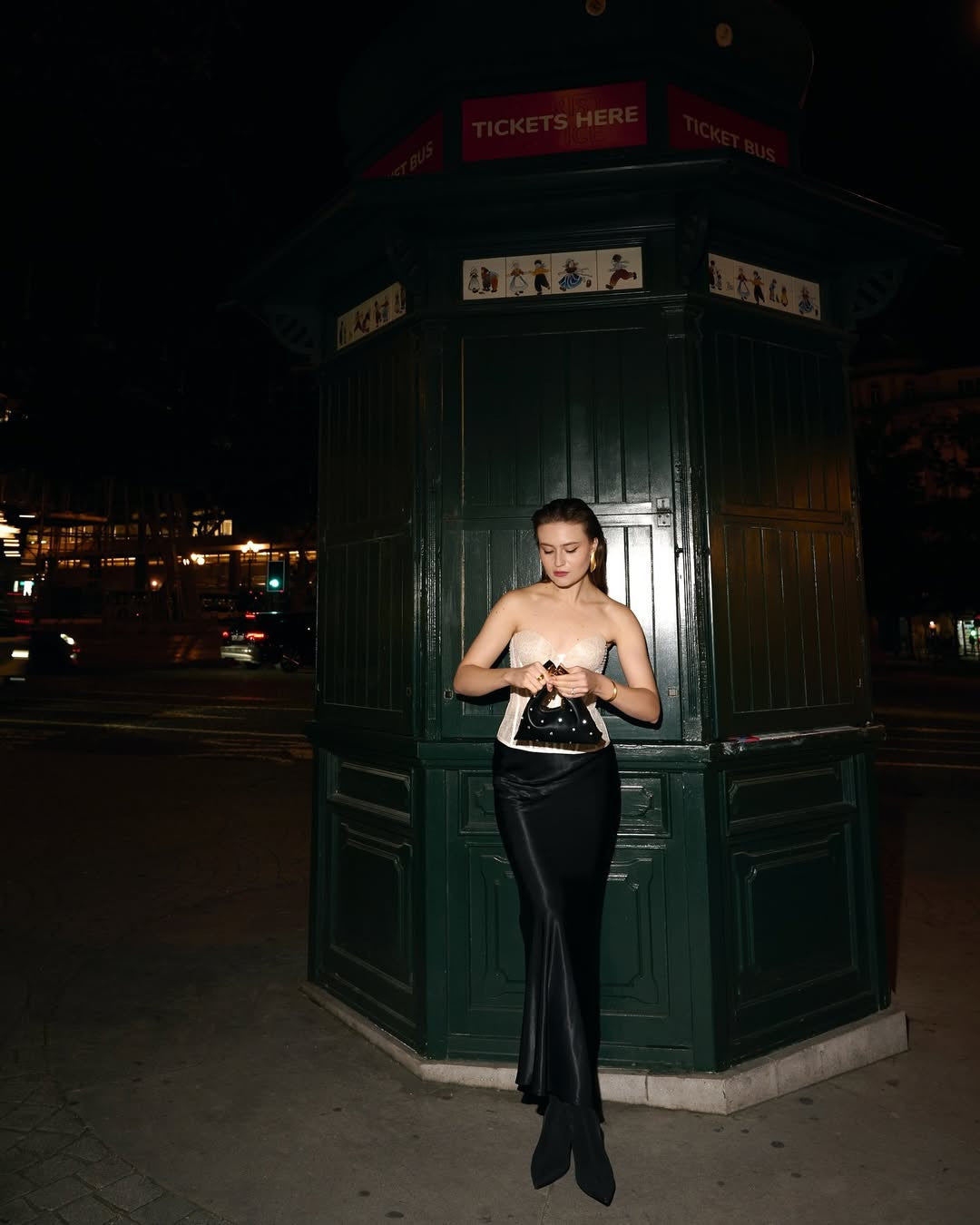 A young woman with dark hair, wearing a strapless white top, a long black skirt, and black boots, stands at night in front of a dark green kiosk with TICKETS HERE and TICKET BUS signs visible at the top. She is holding a small black handbag and appears to be looking down at it. The background is dark with blurry lights, suggesting a city street scene at night.