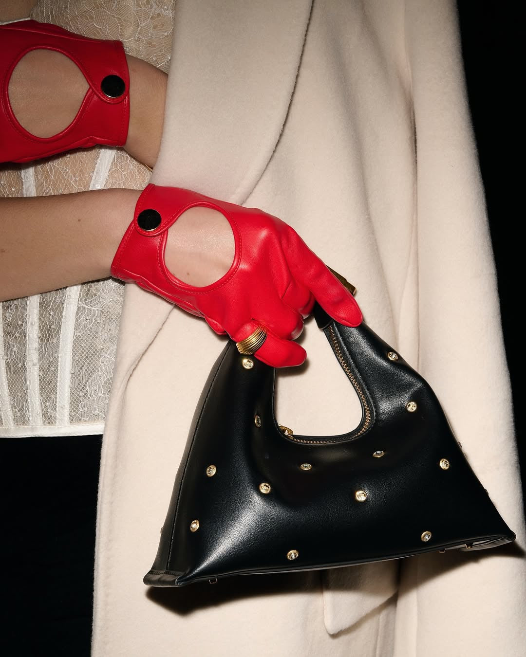 A close-up shot of a person wearing bright red leather gloves with circular cutouts and black button details, holding a black, triangular-shaped handbag adorned with gold studs. The person is also wearing a light-colored, possibly cream or off-white, coat or jacket, and a white, textured garment, possibly lace, is visible underneath the coat. A ring is visible on the gloved hand holding the bag.