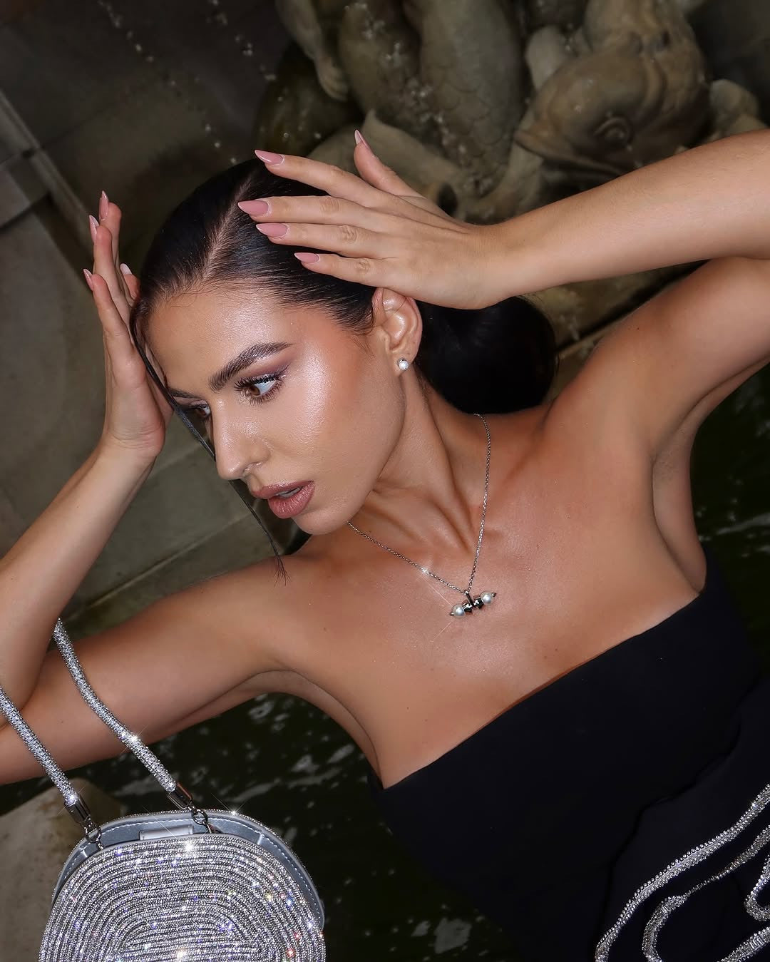 A woman with dark hair pulled back, wearing a black strapless top, a pearl necklace with a pendant, and pearl earrings, holds a shimmering silver handbag while posing near a water feature with a stone sculpture in the background.