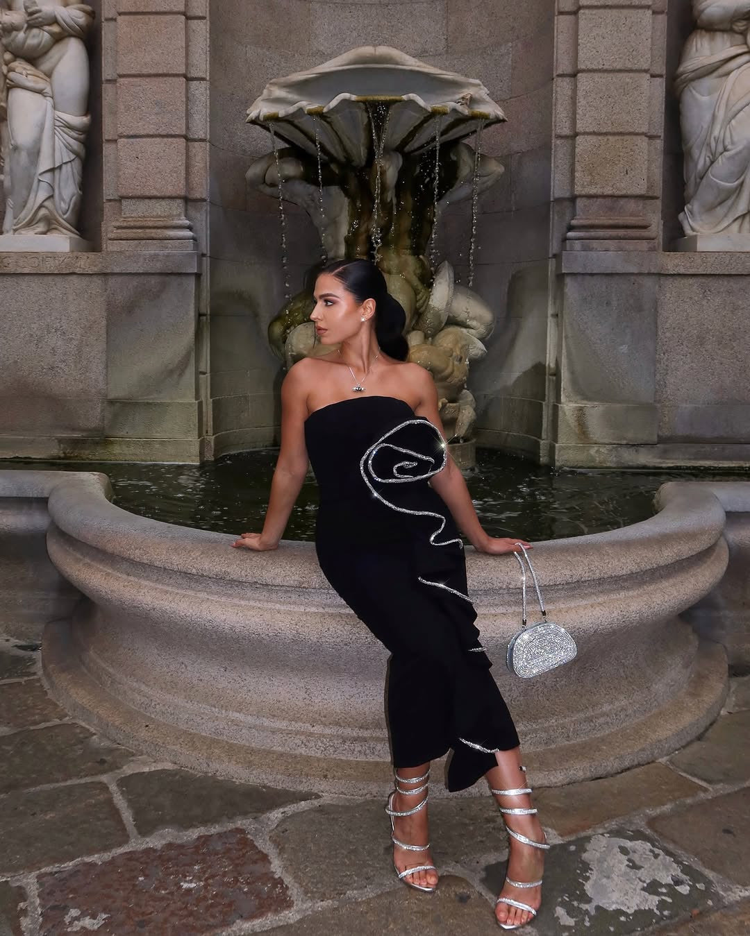 A woman in a black strapless dress with a silver ruffled detail, silver high-heeled sandals, and a sparkling silver clutch bag sits on the edge of a stone fountain. She has dark hair pulled back in a ponytail and is looking to her left. In the background, there is a stone fountain with water flowing and classical statues on either side. The setting appears to be outdoors, possibly in a courtyard or garden with stone paving.