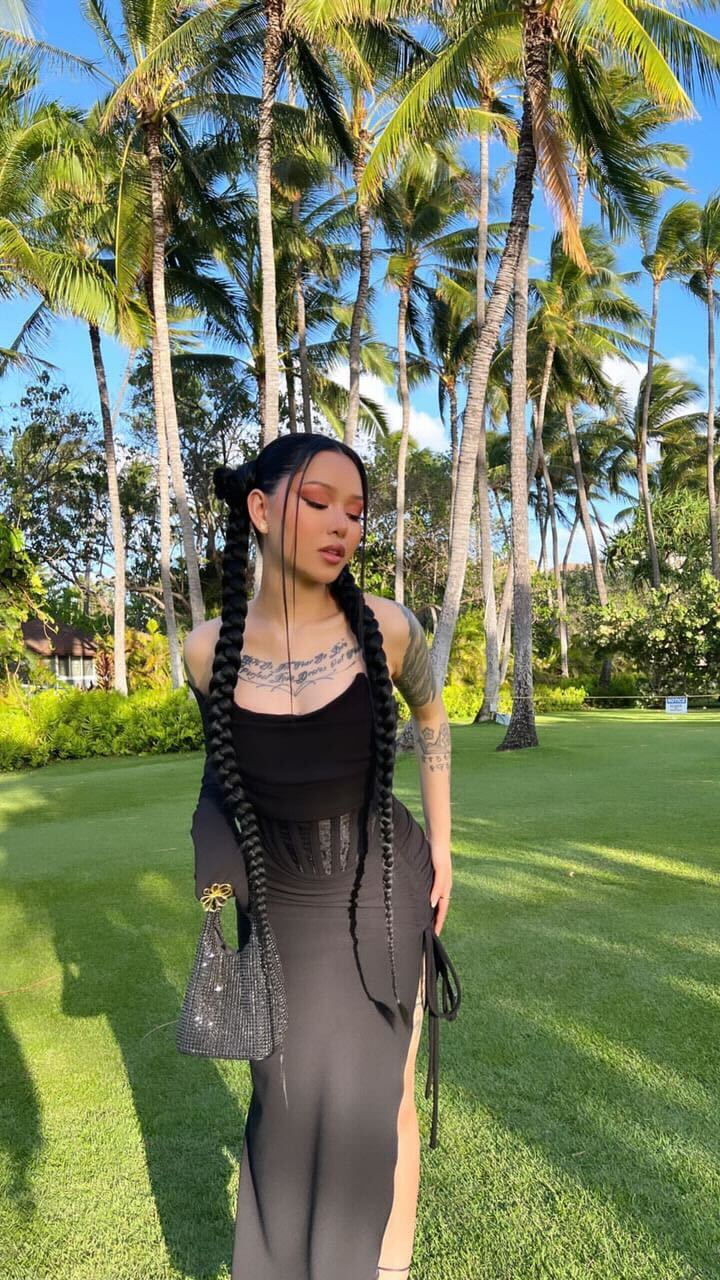 A woman with long, dark braids stands in a grassy area with palm trees in the background, holding a small, embellished handbag.