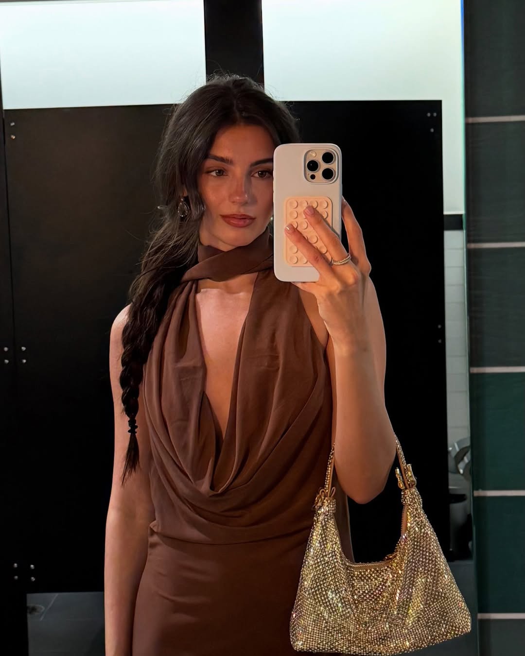 A woman with long, dark braided hair, wearing a brown cowl-neck dress and holding a sparkly gold handbag, takes a mirror selfie in what appears to be a public restroom. 