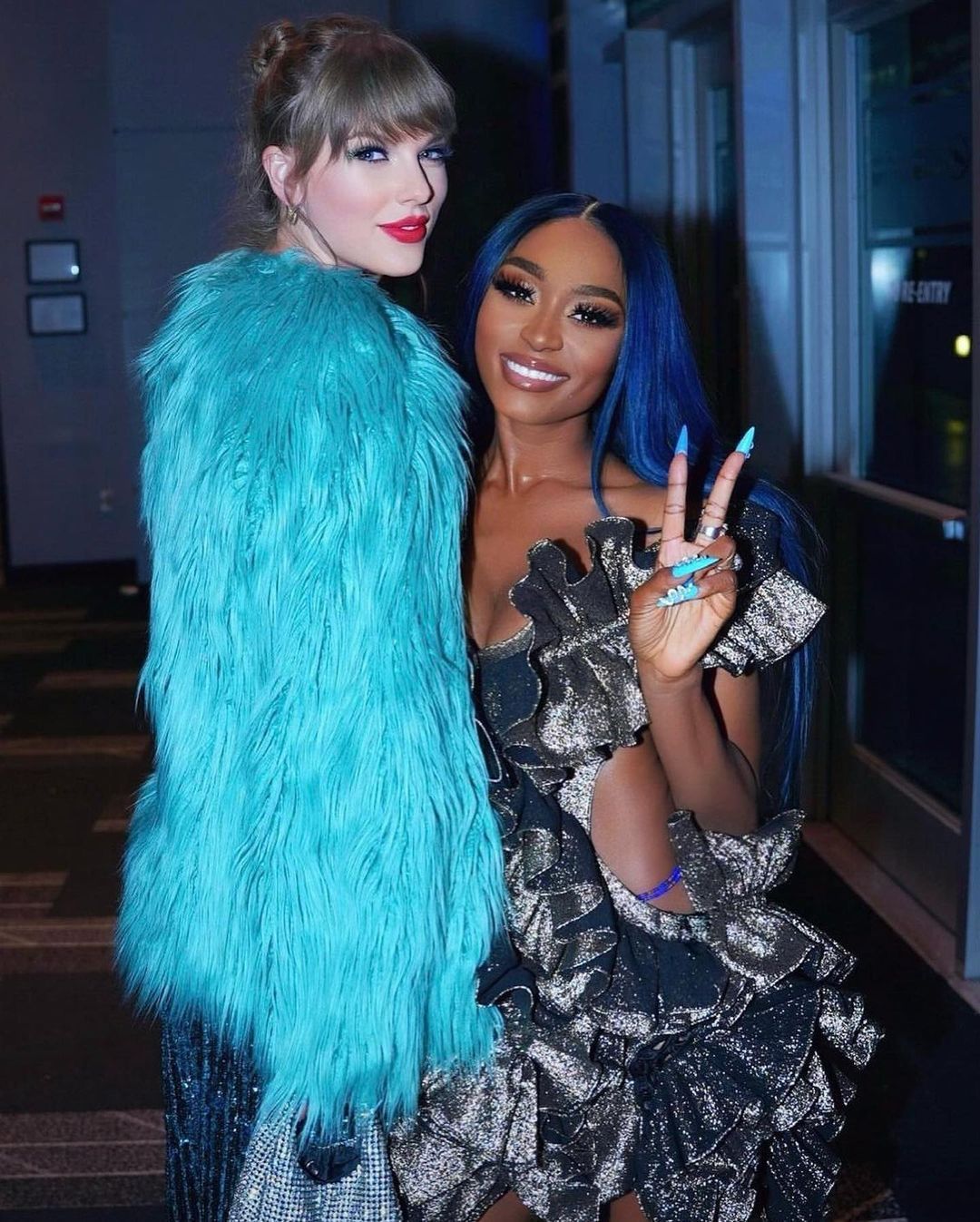 Taylor Swift, wearing a shimmering outfit and a bright turquoise faux fur coat, poses next to Sonyae Elise, who has long blue hair and is wearing a ruffled metallic dress, with Elise making a peace sign gesture.