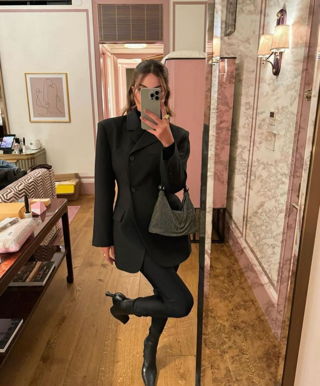 A woman in a black blazer and black pants poses for a mirror selfie in a room, holding a phone to her face and a dark-colored textured handbag.
